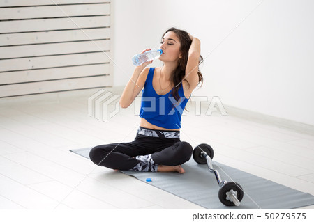 Healthy lifestyle, people and sport concept - Smiling woman sitting on yoga mat and drinking water Healthy lifestyle, people and sport concept - Smiling woman sitting on yoga mat and drinking water 50279575