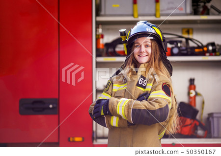 Photo of woman firefighter in helmet with arms crossed standing near fire truck 50350167