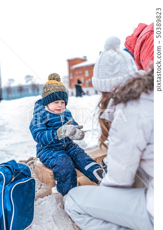 Young mother puts on skates in the winter on a public skating rink. Little boy in a blue jumpsuit 50389823