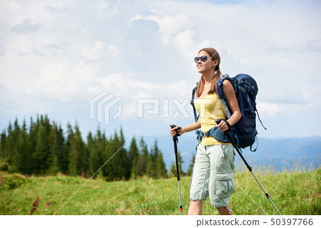 Woman hiker hiking on grassy hill, wearing backpack, using trekking sticks in the mountains 50397766
