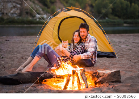 people, summer tourism and nature concept - couple near camp fire warming up seen from the tent 50398566