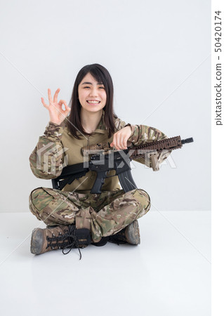 在白色背景上穿著迷彩服裝並且有一把氣槍的女孩也拿著一個圓圈用手指 在白色背景上穿著迷彩服裝並且有一把氣槍的女孩也拿著一個圓圈用手指 50420174