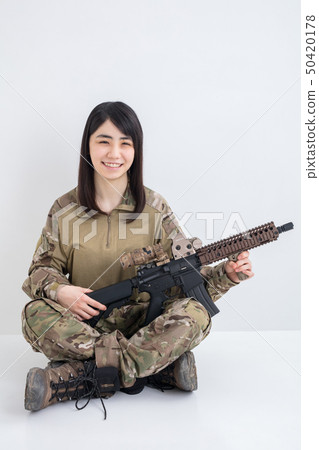 拿著airsoft的一個微笑的女孩穿在白色背景的偽裝成套裝備 拿著airsoft的一個微笑的女孩穿在白色背景的偽裝成套裝備 50420178