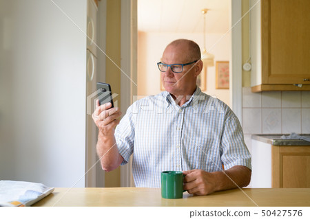 Senior Man Using Phone While Drinking Coffee By The Window Senior Man Using Phone While Drinking Coffee By The Window 50427576