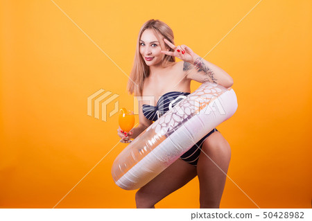 Beautiful in swimming suit showing victory sign and holding orange cocktail over yellow background 50428982