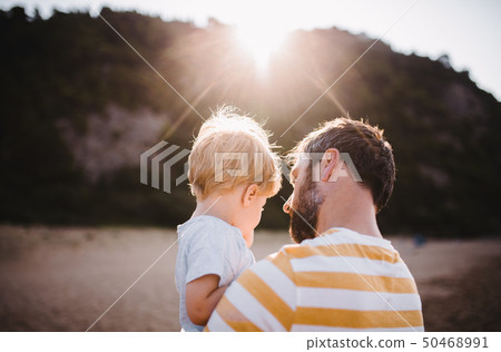 Rear view of father with a toddler boy standing on beach on summer holiday at sunset. 50468991
