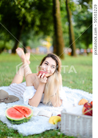 Young female person lying on plaid in park, reading book near watermelon and keeping apple. 50503229