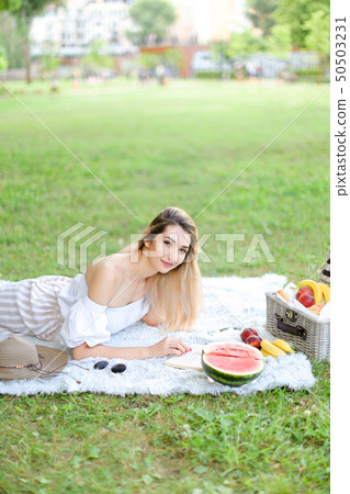 Young pretty girl reading book and lying on plaid near fruits and hat, grass in background. 50503231