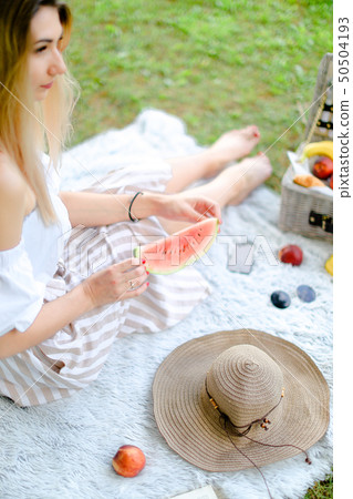 Young blonde woman sitting on plaid near fruits and hat, eating watermelon, grass in background. Young blonde woman sitting on plaid near fruits and hat, eating watermelon, grass in background. 50504193