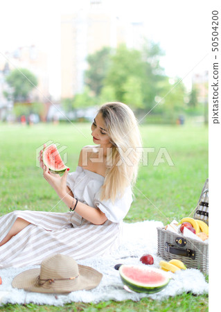 Young girl eating watermelon, sitting on plaid near fruits and hat with grass in background. Young girl eating watermelon, sitting on plaid near fruits and hat with grass in background. 50504200