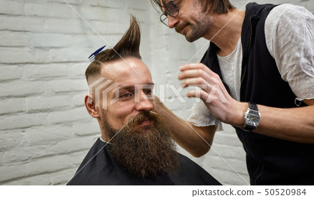 Male barber makes a mohawk hairstyle using clipper of a adult man with beard 50520984