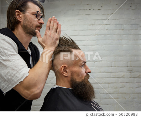 Male barber makes a mohawk hairstyle using clipper of a adult man with beard Male barber makes a mohawk hairstyle using clipper of a adult man with beard 50520988
