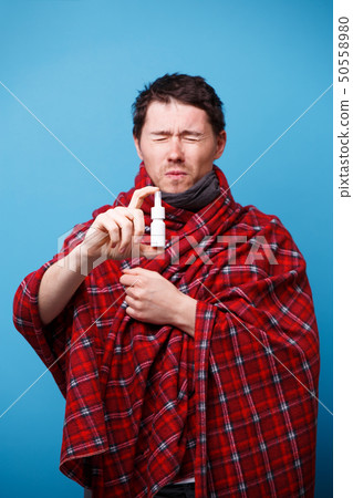 A wrapped in a blanket sick man is standing with nasal spray in his hand 50558980