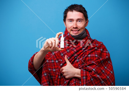 A wrapped in a blanket sick man is standing with nasal spray in his hand A wrapped in a blanket sick man is standing with nasal spray in his hand 50590384