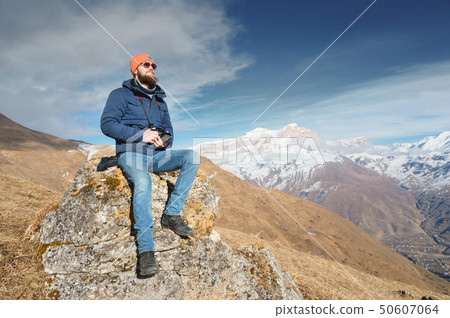 Portrait of a bearded traveler photographer in sunglasses and a cap sits on a rock with mirror Portrait of a bearded traveler photographer in sunglasses and a cap sits on a rock with mirror 50607064