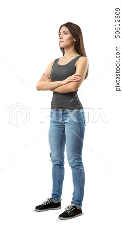 Young woman in gray top and blue jeans standing in half-turn with arms folded, looking up and away 50612809