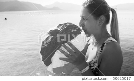 Black and white portrait of beautiful young woman blowing up colorful beach ball at sea 50615017