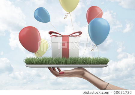 Side view of woman's hand holding digital tablet with green grass on screen, gift box standing on Side view of woman's hand holding digital tablet with green grass on screen, gift box standing on 50621723