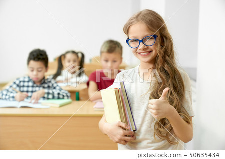 Schoolgirl in classroom holding books, showing thumb up. 50635434