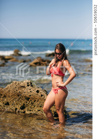 Beautiful young woman with brunette hair in pink swimsuit is posing on the beach near rocks on the Beautiful young woman with brunette hair in pink swimsuit is posing on the beach near rocks on the 50659032