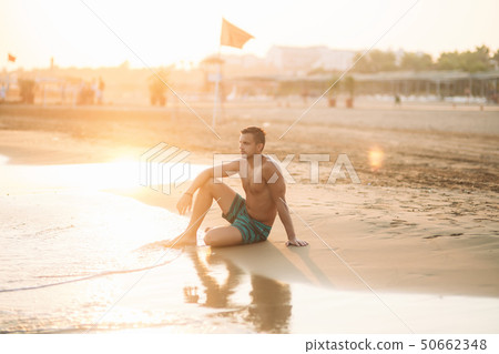 Handsome man on a sandy beach sits on the seashore. Young man on the coast 50662348