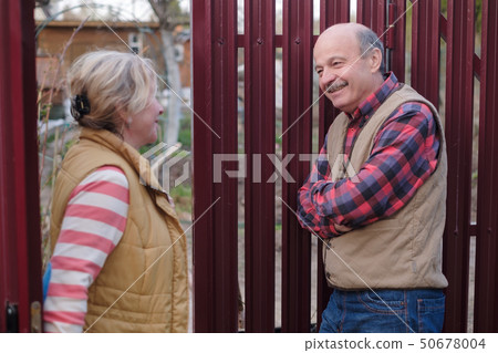 Two senior neighbors takling to each other on sunny day near fence. 50678004