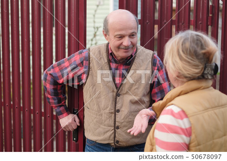 Two senior neighbors takling to each other on sunny day near fence. 50678097
