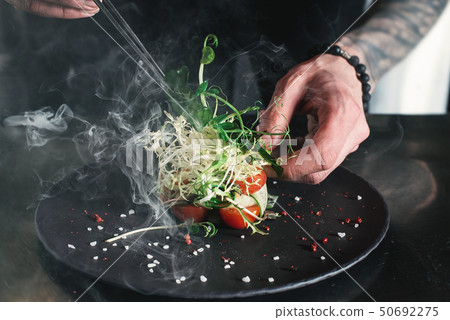 Chef finishing healthy salad on a black plate with tweezers. almost ready to serve it on a table Chef finishing healthy salad on a black plate with tweezers. almost ready to serve it on a table 50692275