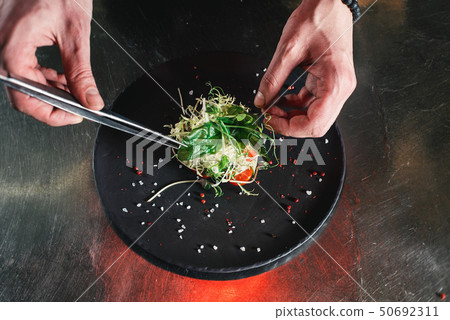 Chef finishing healthy salad on a black plate with tweezers. almost ready to serve it on a table Chef finishing healthy salad on a black plate with tweezers. almost ready to serve it on a table 50692311