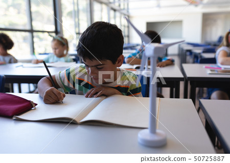 Schoolboy writing on notebook at desk in classroom 50725887