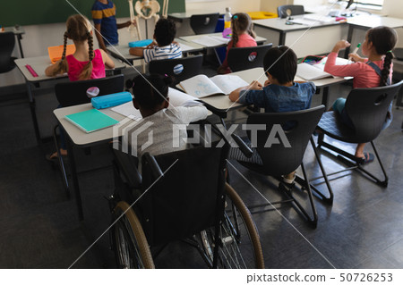 Rear view of disable schoolboy with classmate studying and sitting at desk in classroom Rear view of disable schoolboy with classmate studying and sitting at desk in classroom 50726253