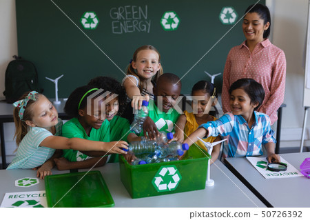 Schoolkids putting bottles in recycle container at desk in classroom 50726392