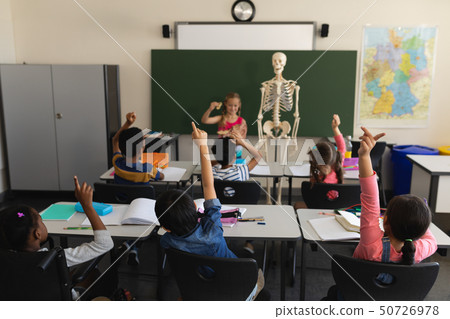 Rear view of schoolkids raising hands and sitting at desk in classroom 50726978