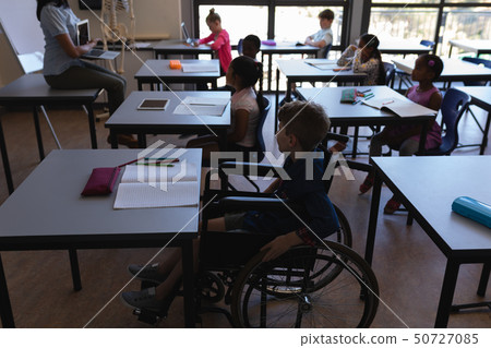 Disable schoolboy with classmates studying and sitting at desk in classroom 50727085