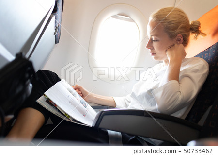 Woman reading in flight magazine on airplane. Female traveler reading seated in passanger cabin. Sun 50733462