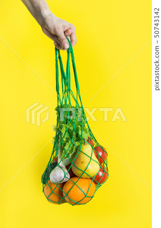 Mesh bag of fruits, vegetables, greens in woman's hand. Zero waste. Plastic free. Yellow background. 50743142