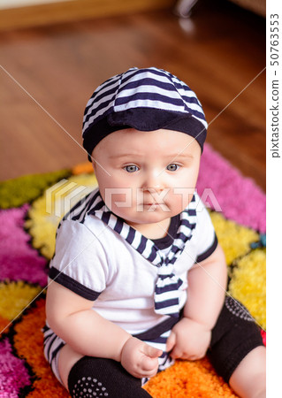A pretty baby in a striped shirt and hats seated on the mat in the room 50763553