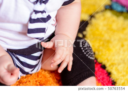 A pretty baby in a striped shirt and hats seated on the mat in the room 50763554