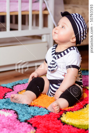 A pretty baby in a striped shirt and hats seated on the mat in the room 50763593