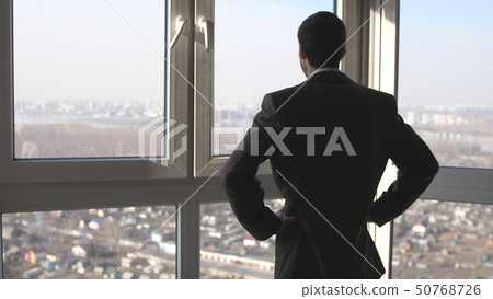 Back view of young successful businessman standing in front of windows looking into distance to the Back view of young successful businessman standing in front of windows looking into distance to the 50768726