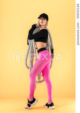 Attractive woman in pink and black sportswear with heavy ropes looking at camera on yellow 50830188