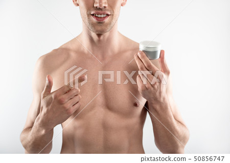 Young man with perfect body holding jar of cosmetic cream and pointing at it Young man with perfect body holding jar of cosmetic cream and pointing at it 50856747