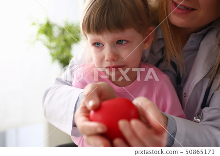 Scared little baby girl visiting doctor holding in hands red toy heart Scared little baby girl visiting doctor holding in hands red toy heart 50865171