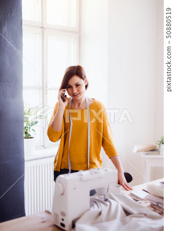 Young creative woman in a studio, using smartphone. A startup of tailoring business. 50880519