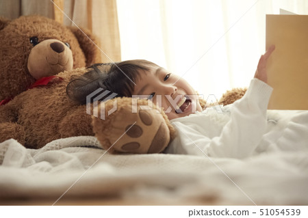Girl reading a picture book on a pillow with a big bear in bed 51054539