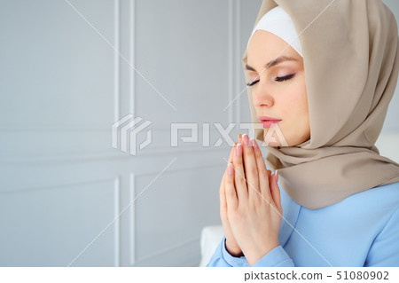 Portrait of young muslim woman in beige hijab and traditional clothes praying with eyes closed 51080902