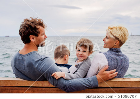 Young family with two small children sitting on bench outdoors on beach. Young family with two small children sitting on bench outdoors on beach. 51086707