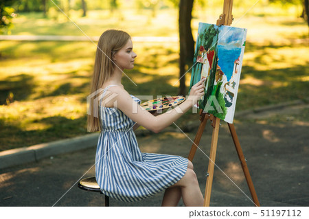 Beautiful blond hair girl sitting on stool and draws a picture in the park using a palette with 51197112