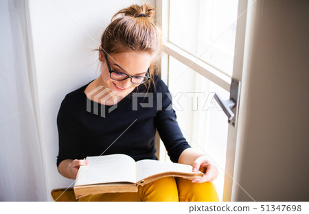 A young female student with book sitting on window sill, studying. 51347698