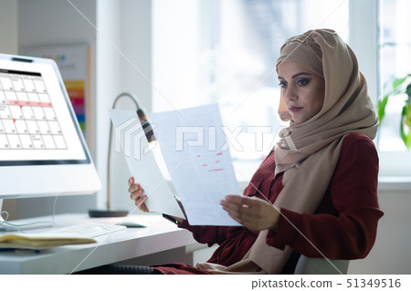 Dark-eyed teacher wearing hijab sitting near computer 51349516